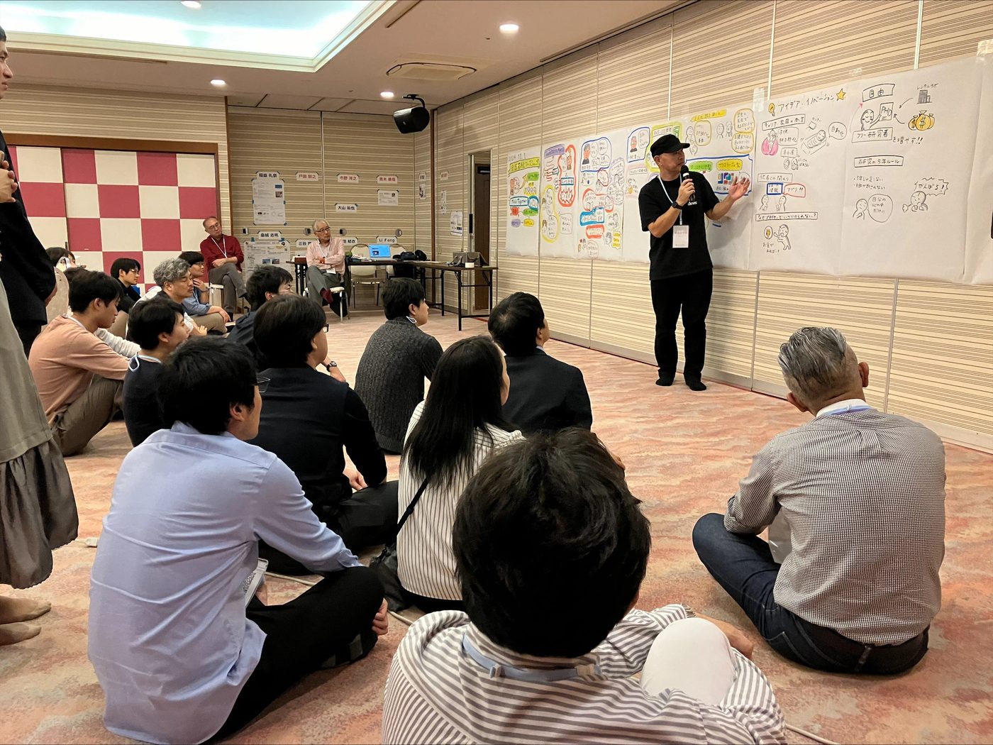 床に座った参加者たちと、グラフィックを指しながら対話する丸山