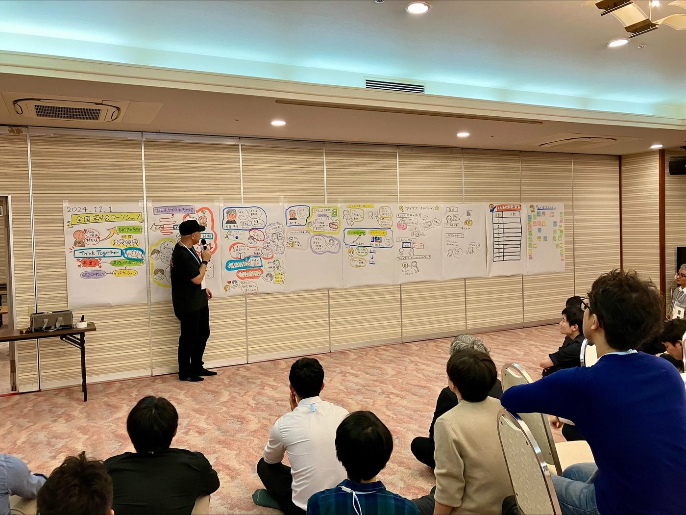 全国若手会ワークショップ。壁一面に並んだグラフィックの前で、参加者に語りかける丸山